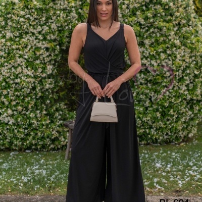 Mulher vestindo macacão preto e segurando mala bege frente a vegetação florida