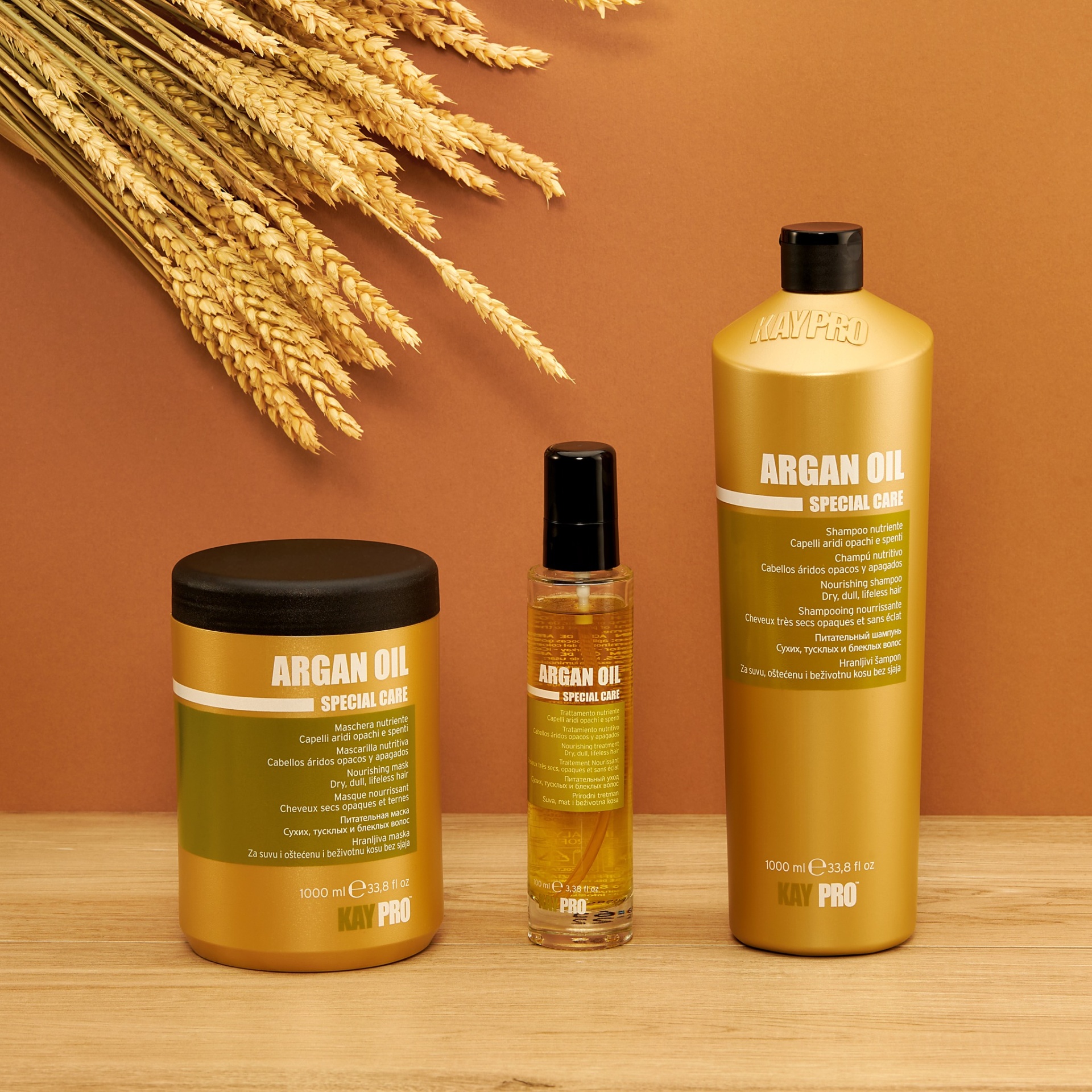 Três produtos dourados e verdes de óleo de argão para cabelo sobre mesa de madeira clara, fundo castanho com espigas secas