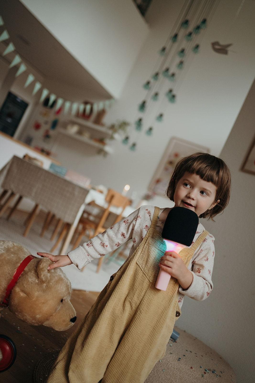 Criança com microfone de brinquedo e urso de peluche numa sala com decoração infantil