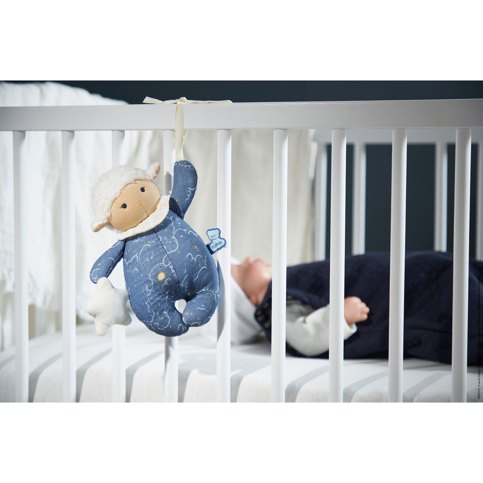 Brinquedo de peluche cordeiro pendurado nas grades de um berço branco com criança deitada ao fundo