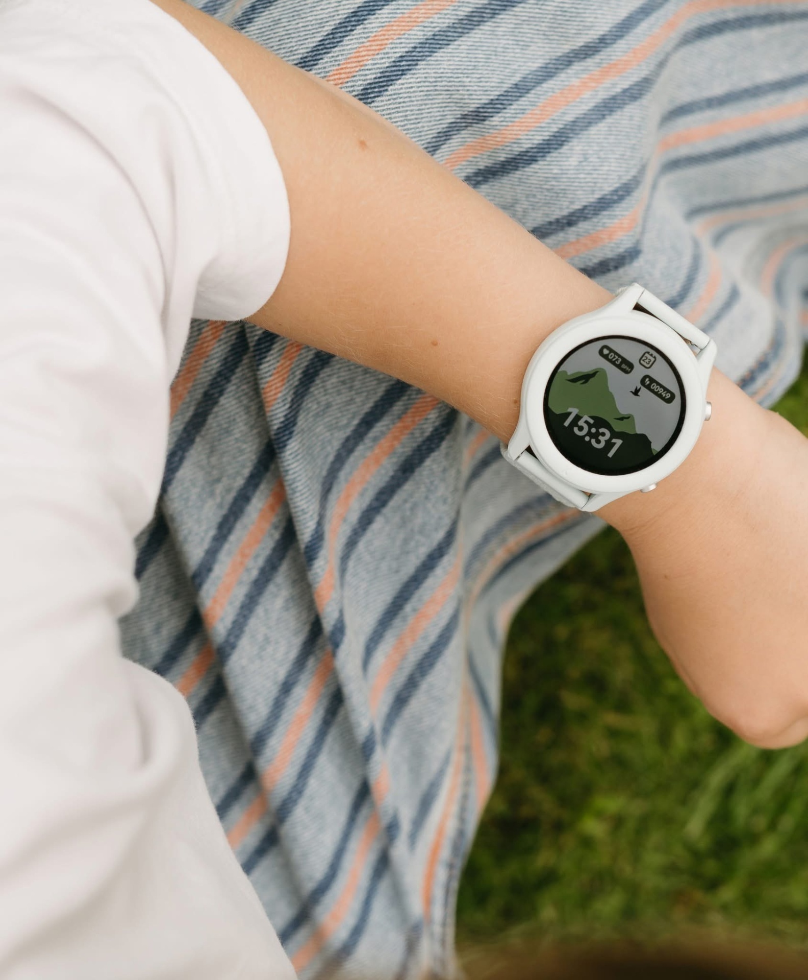 Relógio smartwatch branco com ecrã digital a mostrar 15:31, gráficos e ícones, braço com camisola branca e manta às riscas no fundo.