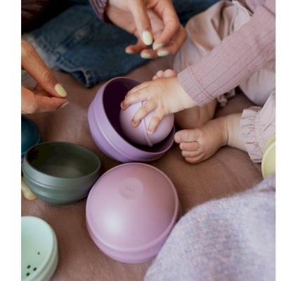 Mãos de adulto e criança a brincar com bolas e tigelas coloridas sobre superfície rosa