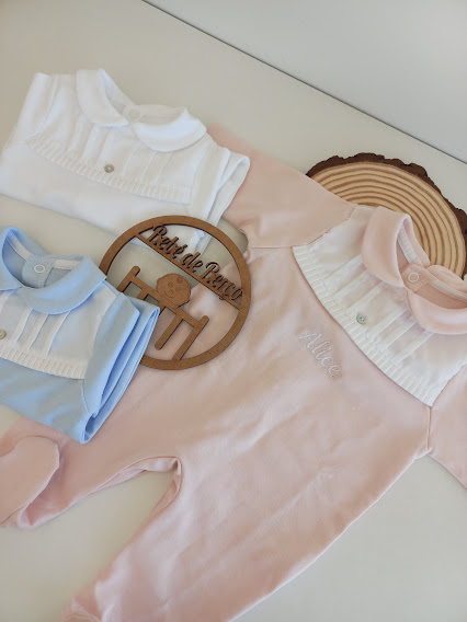 Três macacões para bebé nas cores azul, branco e rosa com detalhes e um ornamento de madeira