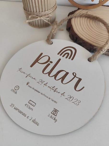 Placa branca redonda com texto castanho sobre nascimento de bebé e fundo com objetos de madeira.