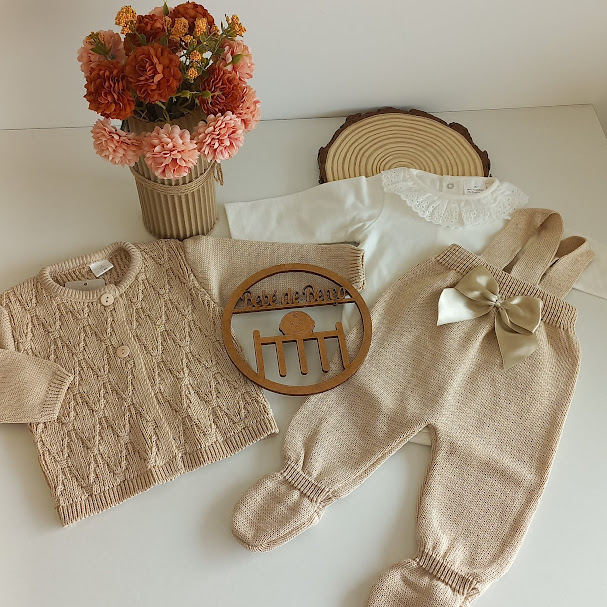 Conjunto de roupa de bebé bege e branco com camisola, body e calças, decorado com flores artificiais e placa de madeira.