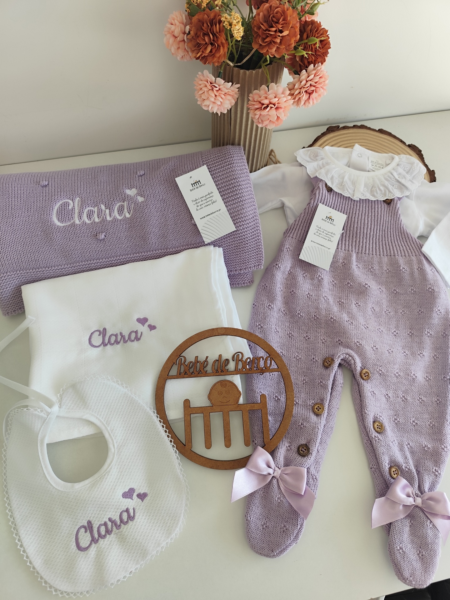Conjunto bebé com manta, babete, toalha bordada e macacão lilás, e decoração de madeira
