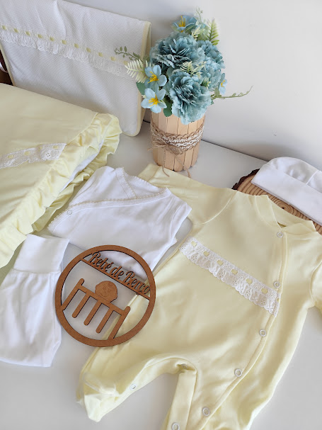 Conjunto de roupa de bebé com babygrow amarelo, body branco, calças brancas, touca branca, almofada amarela, flores azuis e símbolo em madeira com texto