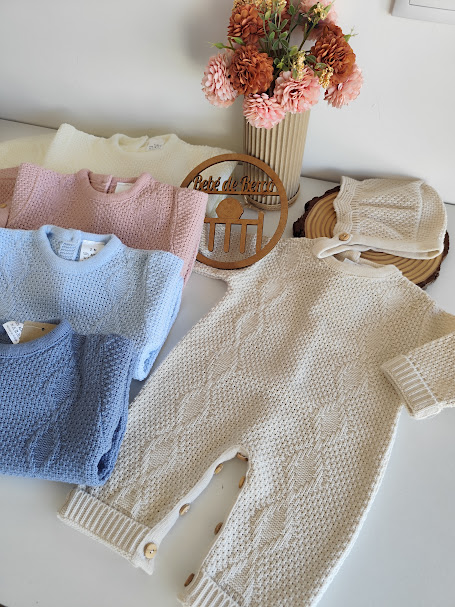 Roupas de bebé em malha de várias cores dispostas sobre uma mesa branca com flores decorativas