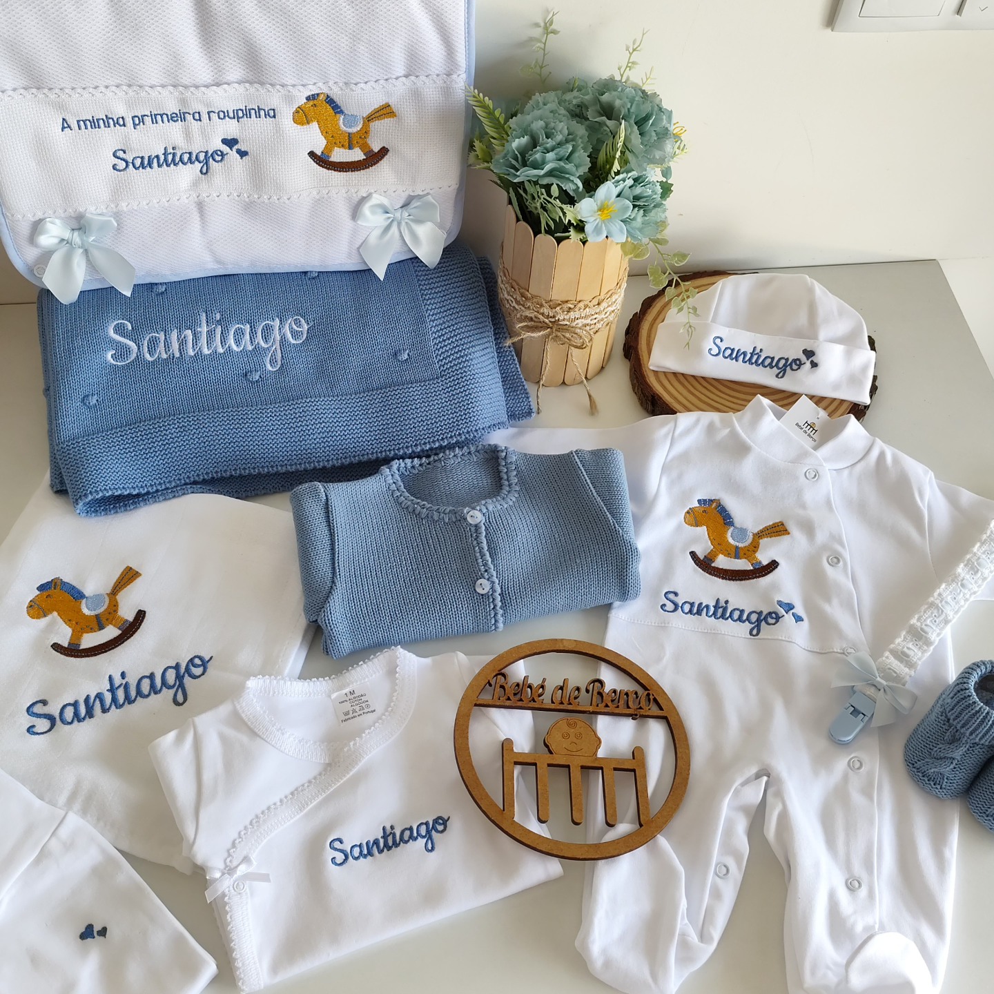 Roupas de bebé brancas e azuis bordadas com nome Santiago e cavalo de baloiço, sobre mesa com decoração floral