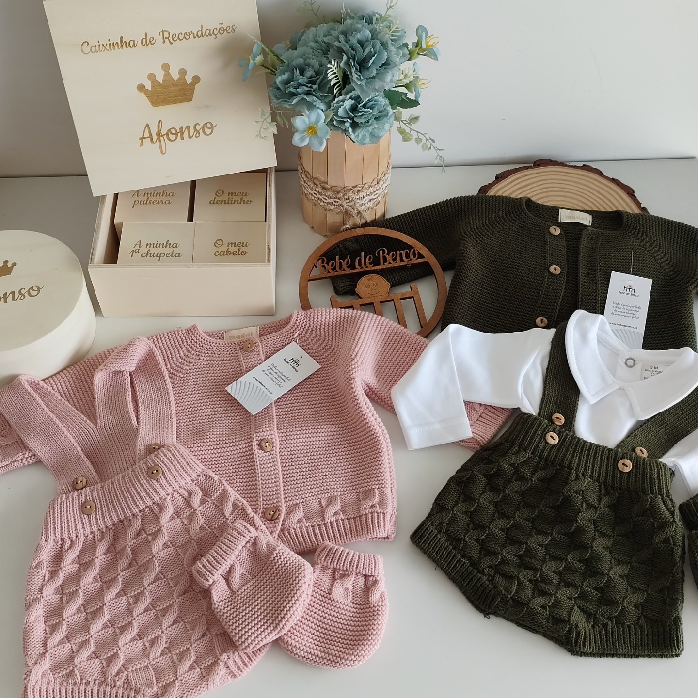Roupas de bebé em tricô rosa e verde com caixa de recordações e vasos de flores azuis