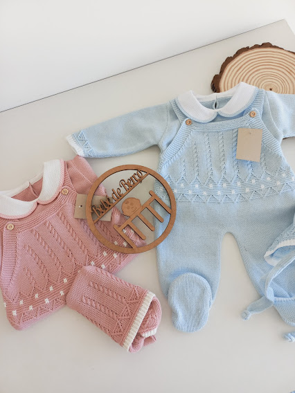 Fatos de bebé em malha rosa e azul claro com gola branca e botões sobre superfície clara com ornamento de madeira.