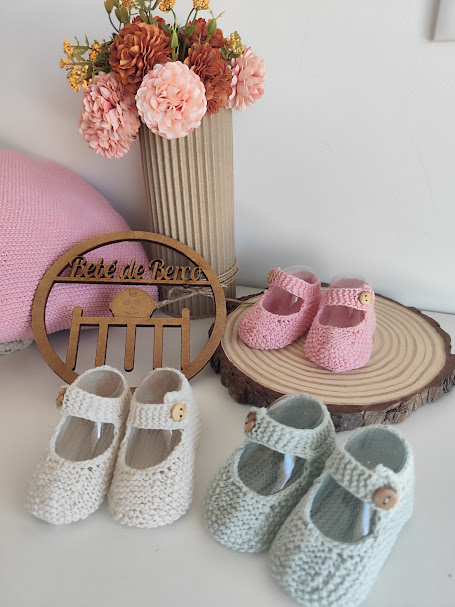 Sapatos de bebé em malha rosa, branca e azul claro com botão de madeira, decoração com flores, almofada rosa e peça em madeira com texto