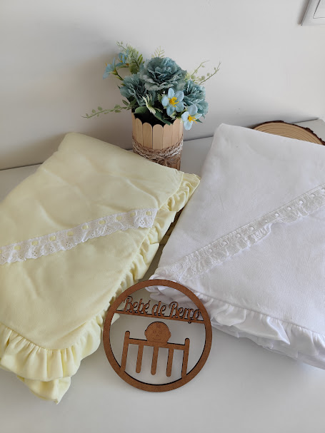 Dois lençóis de berço com folhos e renda, vaso com flores azuis e ornamento de madeira com texto