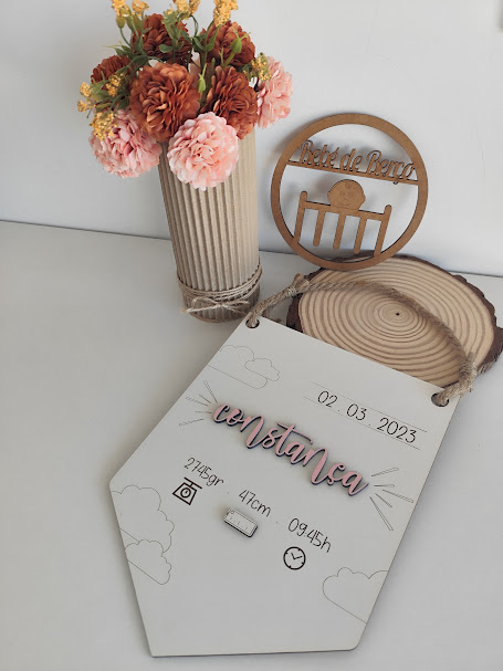 cartaz decorativo com data e nome Constança ao lado de vaso com flores e base redonda de madeira