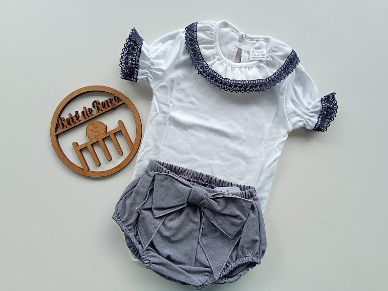 Conjunto de roupa de bebé com t-shirt branca e calções cinzentos e um círculo de madeira com texto em fundo branco.