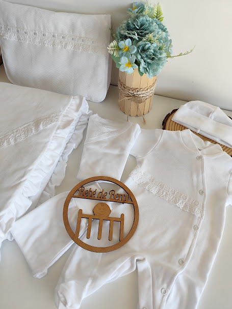 Conjunto branco de roupa de bebé com detalhes rendados e placa de madeira com texto Pé de Roupa