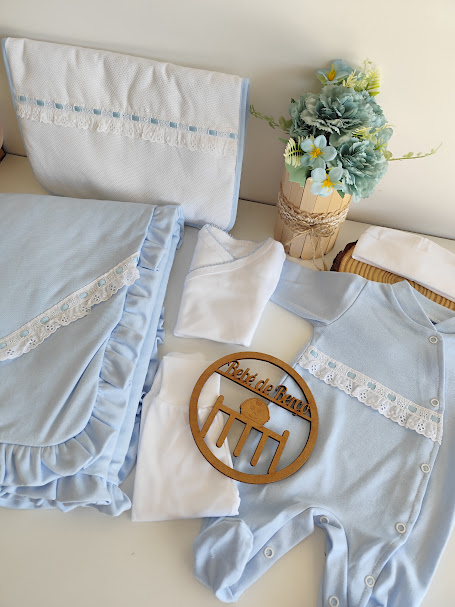 Conjunto de roupa de bebé azul claro e branco com manta, babygrow, fralda, toalha e bolsa de transporte ao lado de um arranjo floral