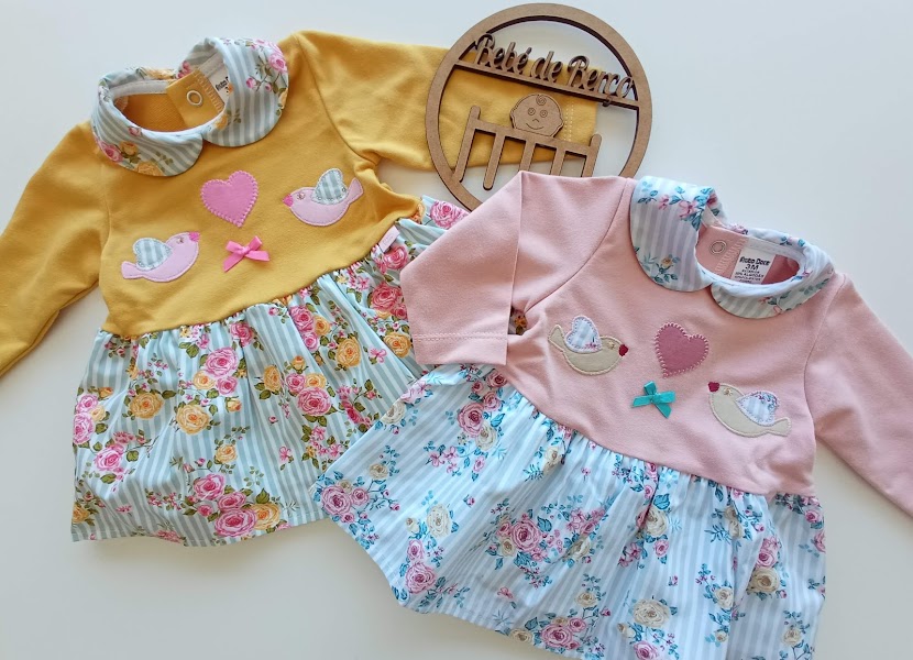 Dois vestidos de bebé coloridos com padrão floral e aplicações decorativas, dispostos ao lado de placa de madeira redonda com texto