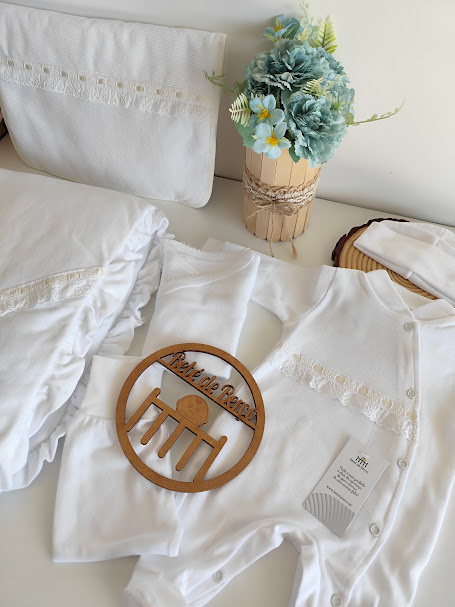 Conjunto de roupa de bebé branco com renda e vaso de flores azuis num ambiente claro