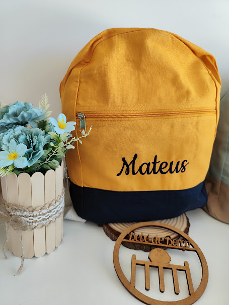 mochila amarela e azul escuro com nome Mateus bordado, ao lado de suporte de madeira com flores artificiais