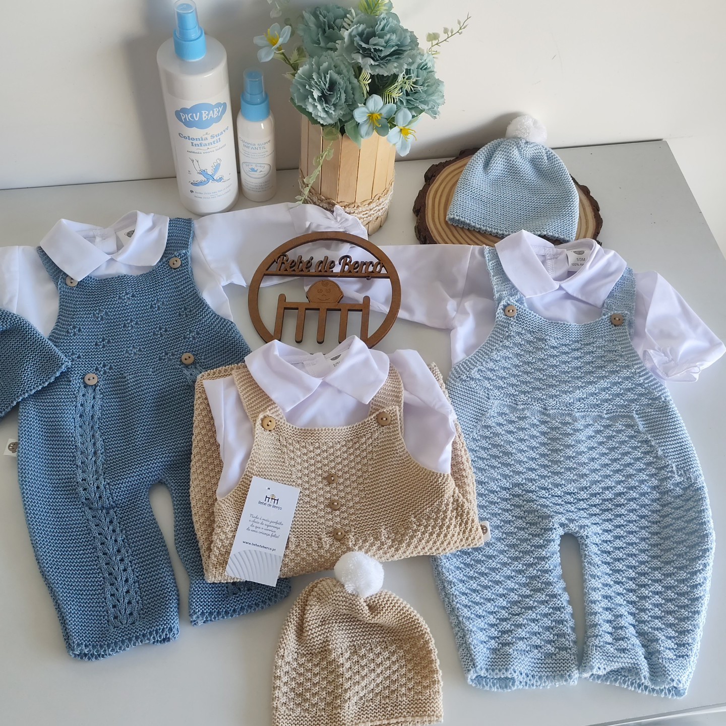 Conjuntos de roupa para bebé com jardineiras de malha azul e bege, camisolas brancas e gorros, com sprays ao fundo
