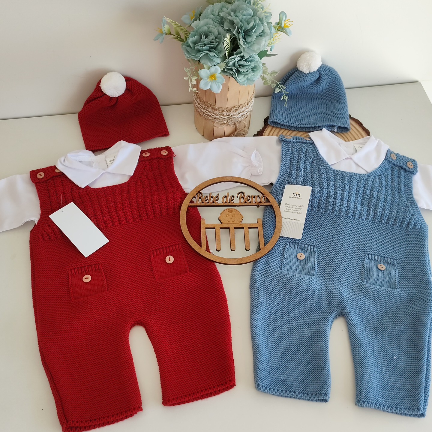 Dois macacões de malha para bebé, vermelho e azul, com gorros de pompom e camisas brancas sobre uma superfície branca com um vaso de flores