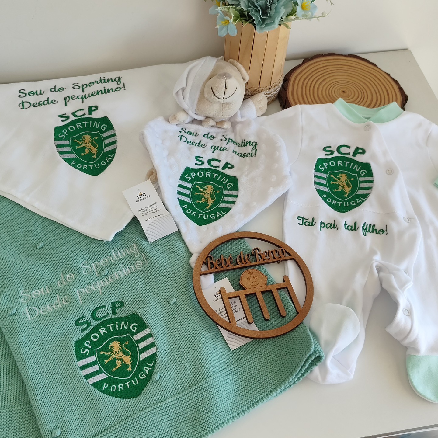 Conjunto de artigos de bebé branco e verde com o emblema do Sporting Portugal e texto em português, junto a peluche e decorativos de madeira