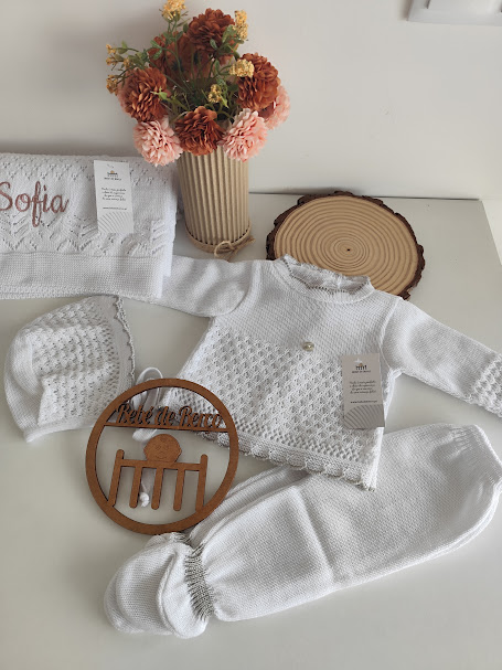Conjunto de roupa de bebé branca em malha, toalha bordada, vaso com flores e decoração de madeira