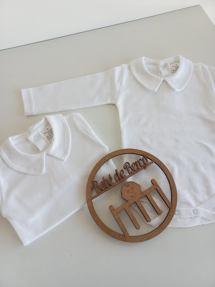Peças de roupa de bebé brancas com gola Peter Pan e placa de madeira com texto Bebe de Boneca