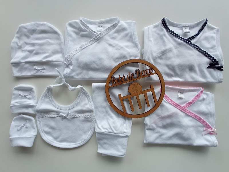 Conjunto de roupa de bebé branca com babete, camisolas com bordas coloridas e acessório de madeira com texto