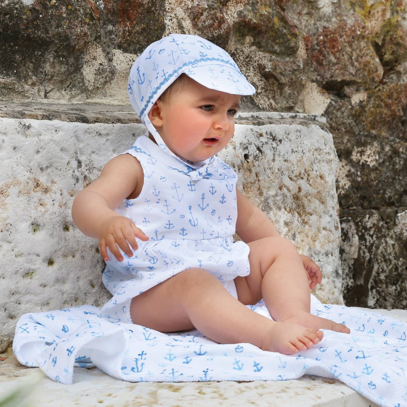 Bebé com roupa branca decorada com âncoras azuis sentado em pedra branca junto a uma parede rústica.
