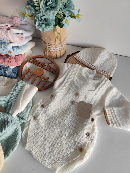 Conjunto de bebé em tricô branco com botões castanhos e gorro, roupa de bebé colorida e flores em vaso sobre superfície branca