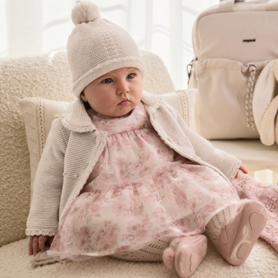 bebé com vestido rosa floral, casaco e gorro bege, sentado num sofá creme com mochila bege ao fundo