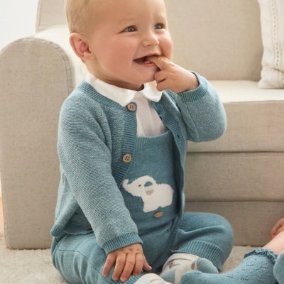 Bebé com fato de malha azul com coelho, camisola com botões e meias azuis sentado em tapete branco perto de sofá bege.