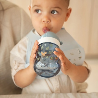 Copo infantil azul com tampa e bico azul claro decorado com animais e folhas, segurado por criança com babete azul.