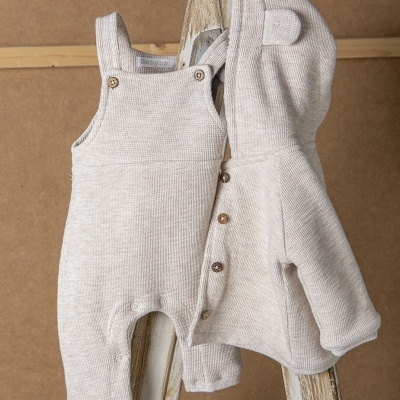 Conjunto de bebé bege claro em malha com jardineiras e casaco com capuz que tem orelhas, pendurado num cabide de madeira