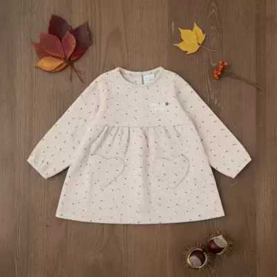 Vestido infantil bege de manga comprida com bolsinhos de coração sobre madeira e folhas de outono.