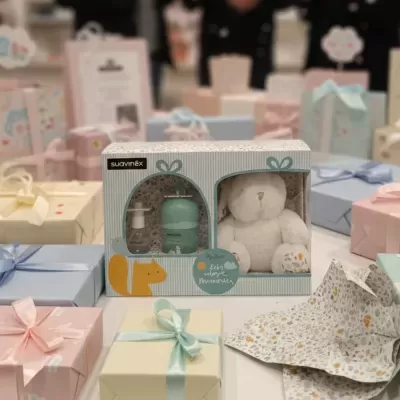 Caixa de presente Suavinex com ursinho de peluche e frascos, rodeada por caixas de presente coloridas.