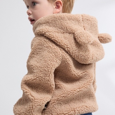 Criança vestindo casaco bege de pelúcia com capuz e orelhas, e calças de ganga azuis