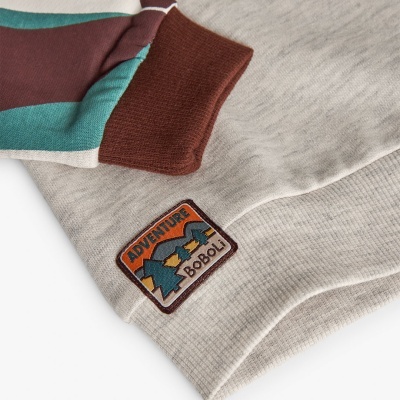 Peça de roupa de malha bege com etiqueta bordada e manga com riscas castanhas e verde-azuladas