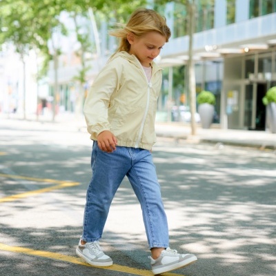 Criança a caminhar na rua com casaco amarelo, calças de ganga e sapatilhas brancas.