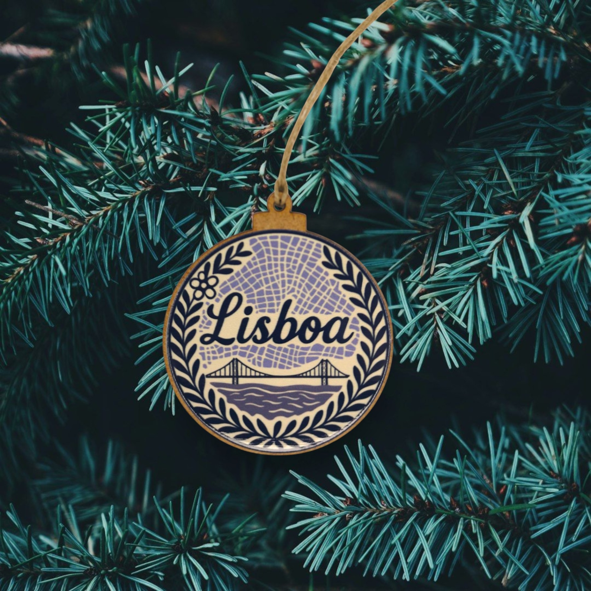 Bola de Natal "Lisboa" Enfeite de Natal redondo em madeira com desenho azul e texto Lisboa pendurado num ramo de pinheiro