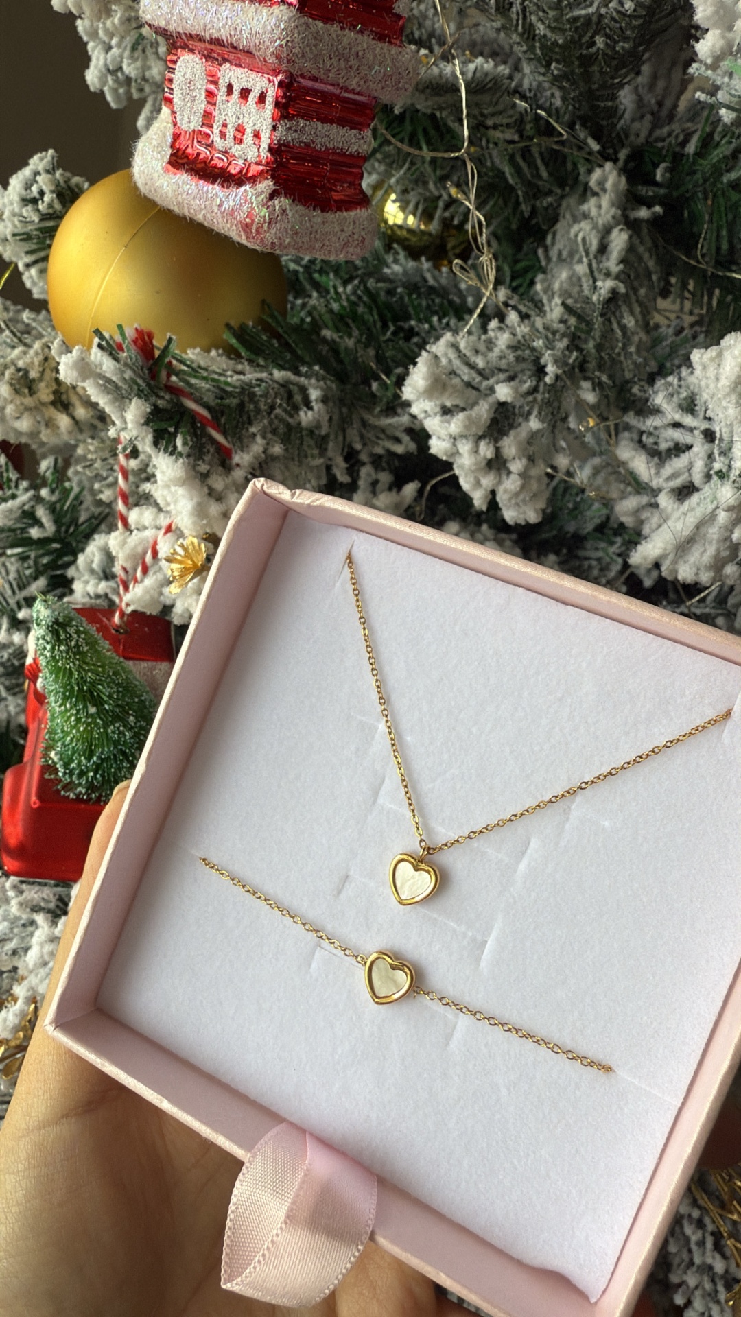 Conjunto de joias douradas com coração numa caixa rosa sobre fundo de árvore de Natal decorada.