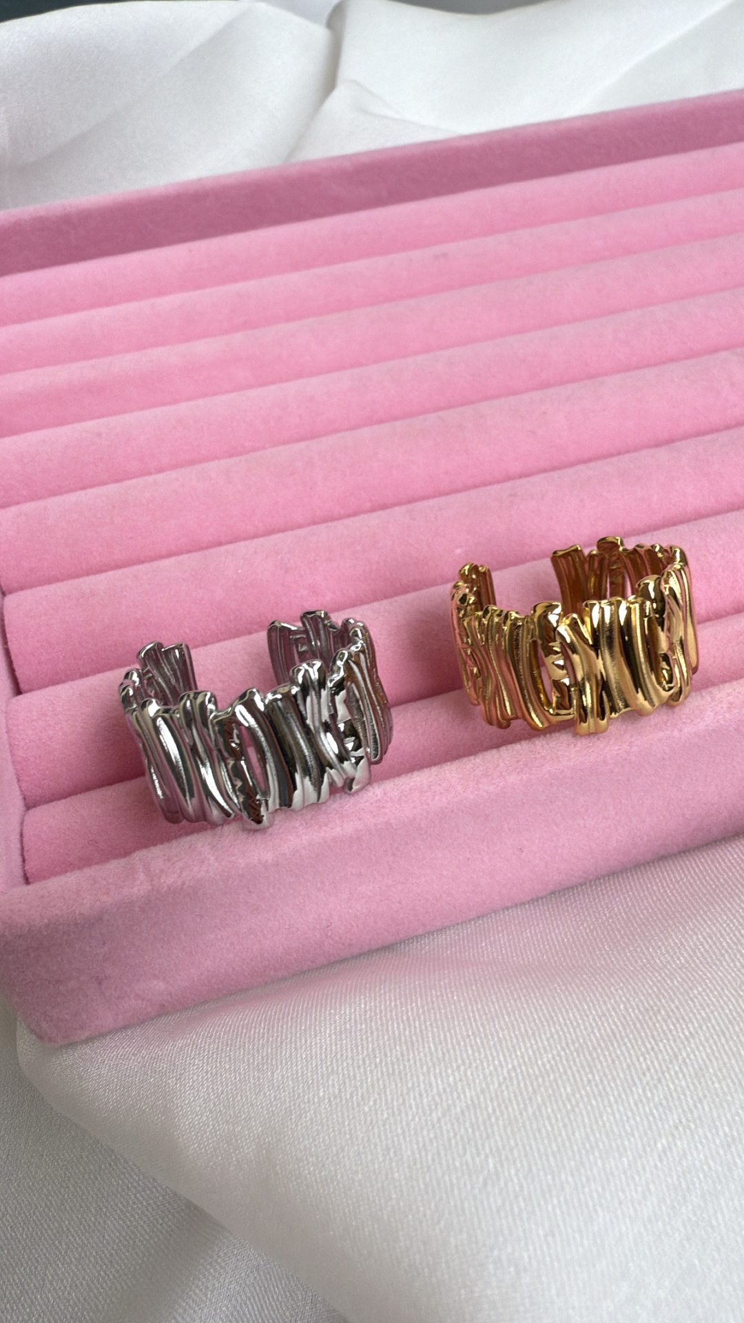 Dois anéis metálicos, um prateado e outro dourado, em caixa rosa com tecido branco.
