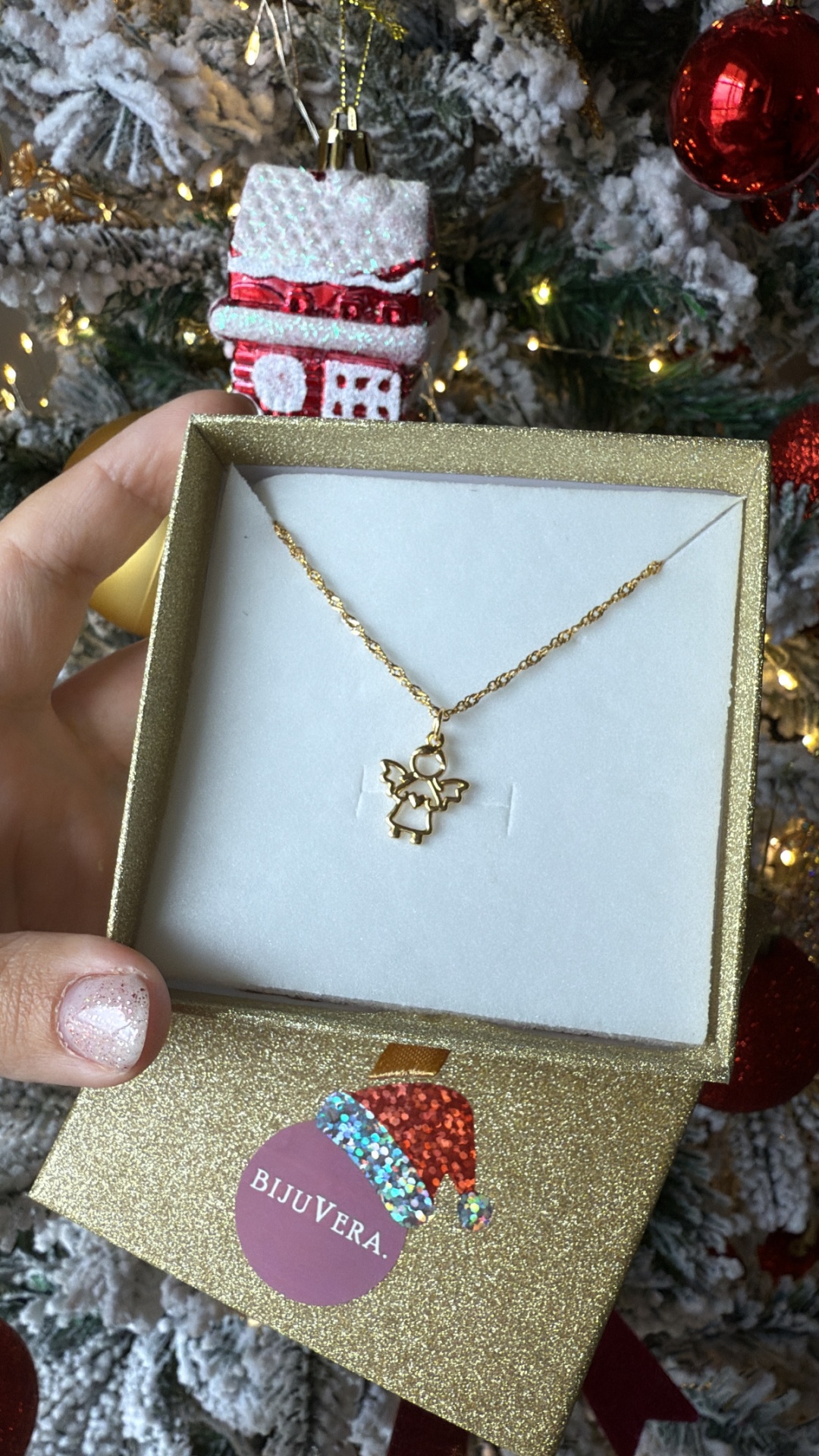 Colar dourado com pendente de anjo numa caixa dourada com etiqueta BIJUVERA frente a uma árvore de Natal decorada