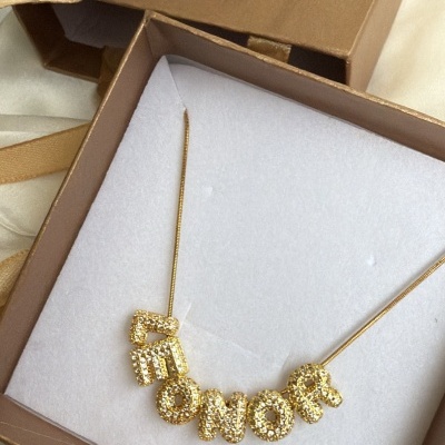 Colar dourado com letras cravejadas dentro de caixa castanha com selo BIJUVERA