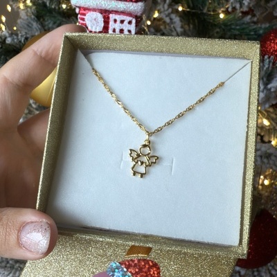 Colar dourado com pendente de anjo numa caixa dourada com etiqueta BIJUVERA frente a uma árvore de Natal decorada