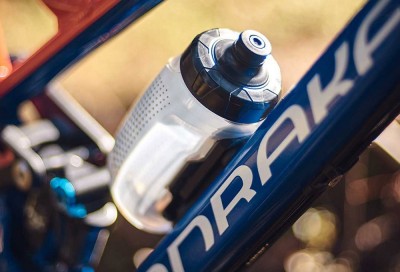 Garrafa de água transparente em suporte preto numa bicicleta azul com texto 'DRAK'