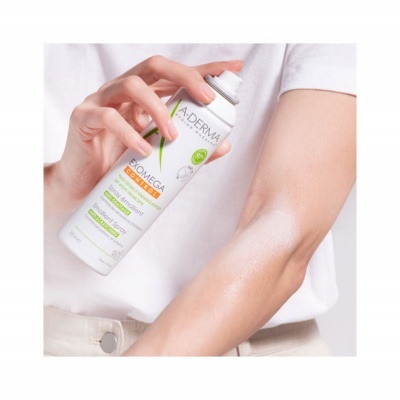 Spray corporal A-DERMA Exomega Control em embalagem branca com detalhes verdes e laranja sendo aplicado no braço