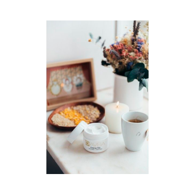 Creme facial em pote branco com etiqueta dourada, vela acesa, chávena branca, prato com grãos e flores secas num fundo branco